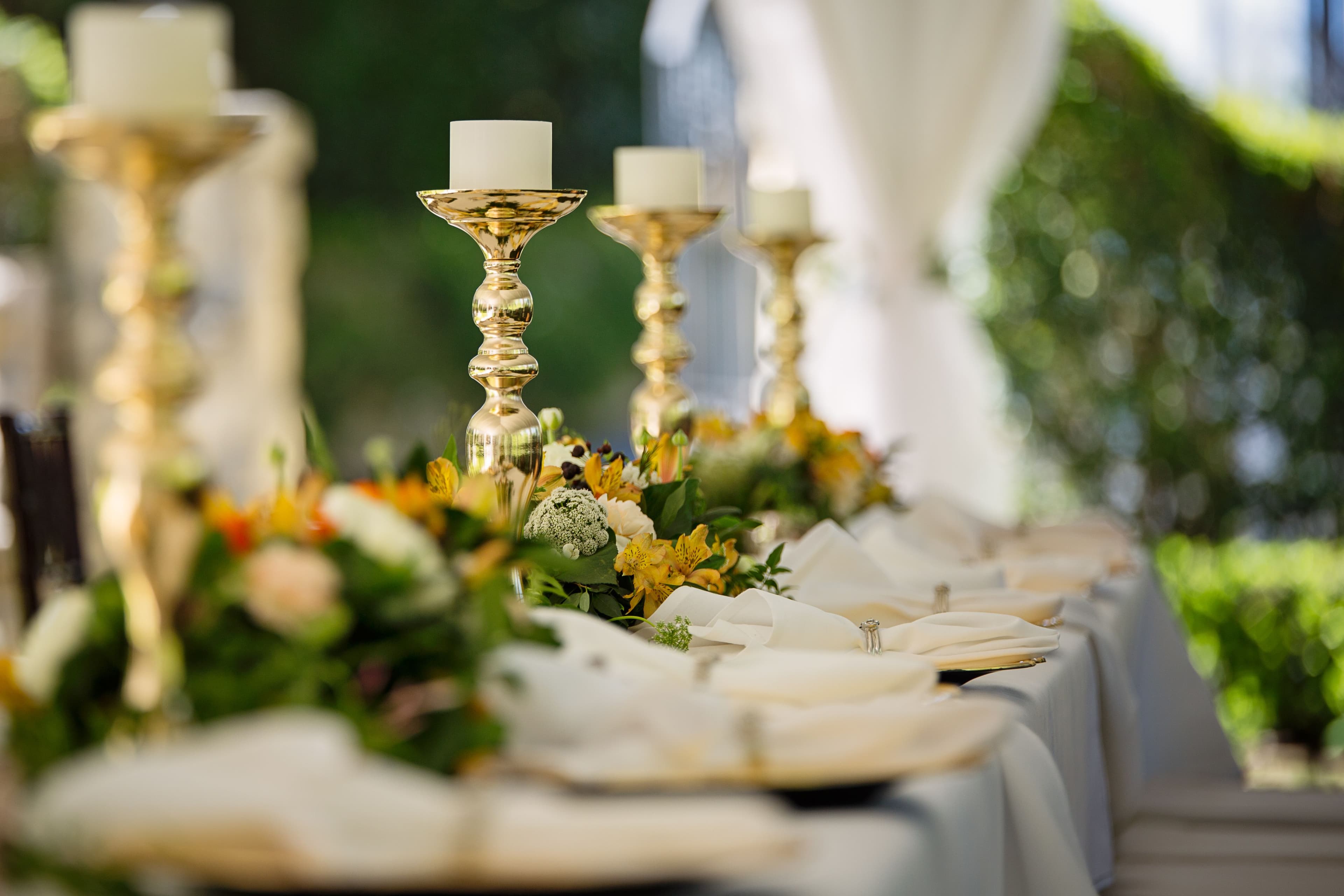 Elegant outdoor wedding table set with gold candlesticks, yellow floral arrangements, and white linens.